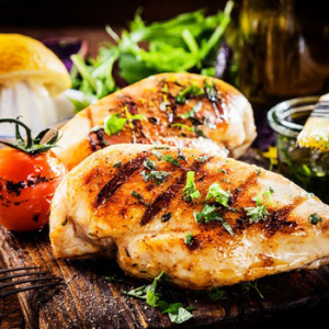 Two chicken breasts with grill marks on them on a wood cutting board with herbs and vegetables in the background.